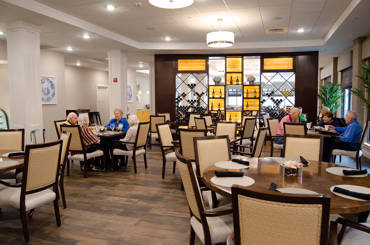 Residents dining and chatting at tables in a bright, well-appointed dining room with wooden floors and a backlit wine display.