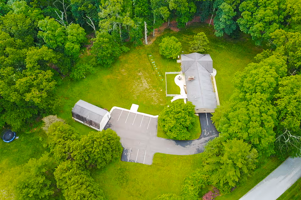 Aerial view of a single-story building with a parking area and small outbuilding surrounded by trees and grassy lawn.