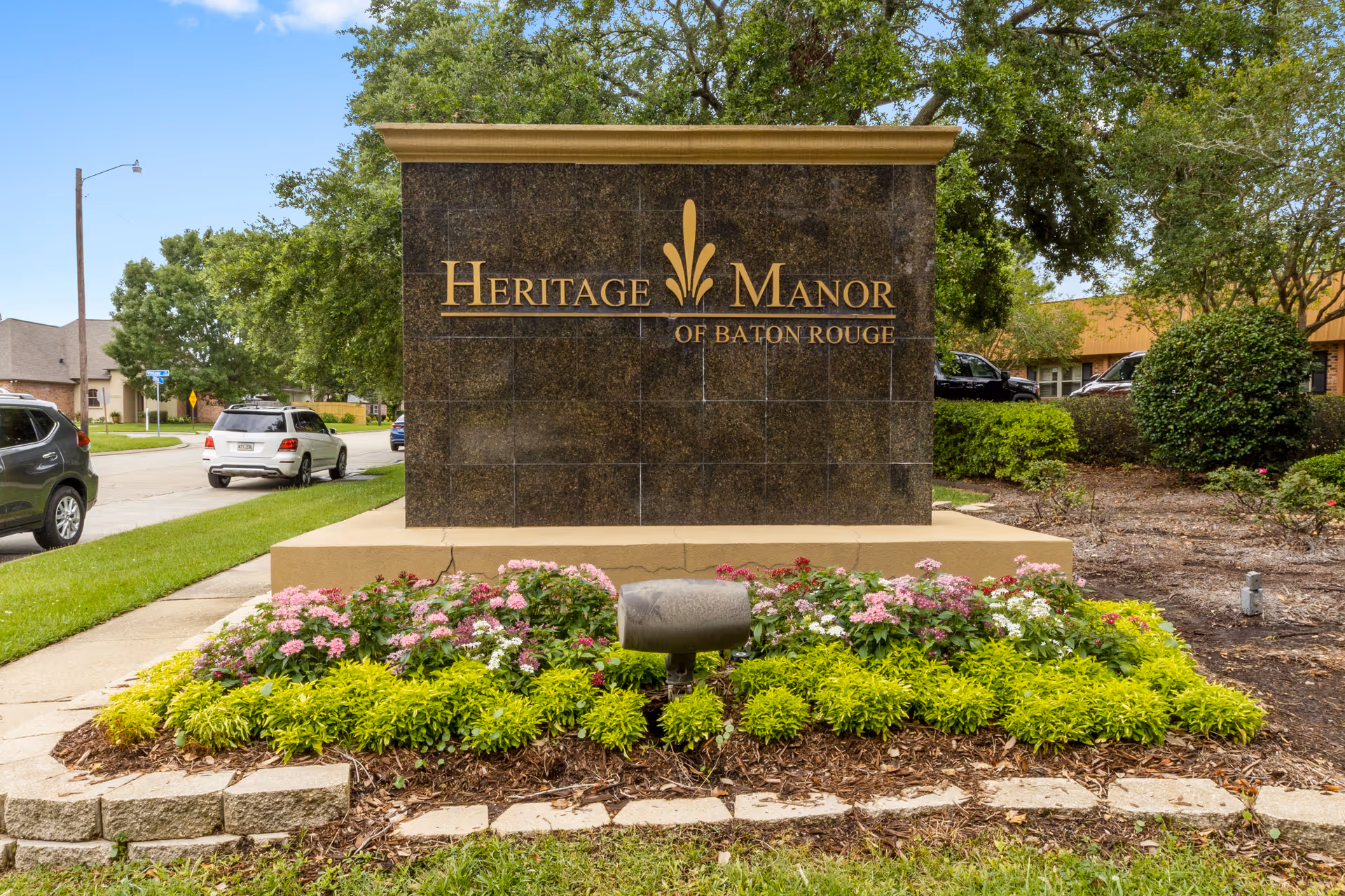 Outdoor stone sign for Heritage Manor of Baton Rouge surrounded by green shrubs and pink flowers, with a street and parked cars visible in the background under a clear blue sky.