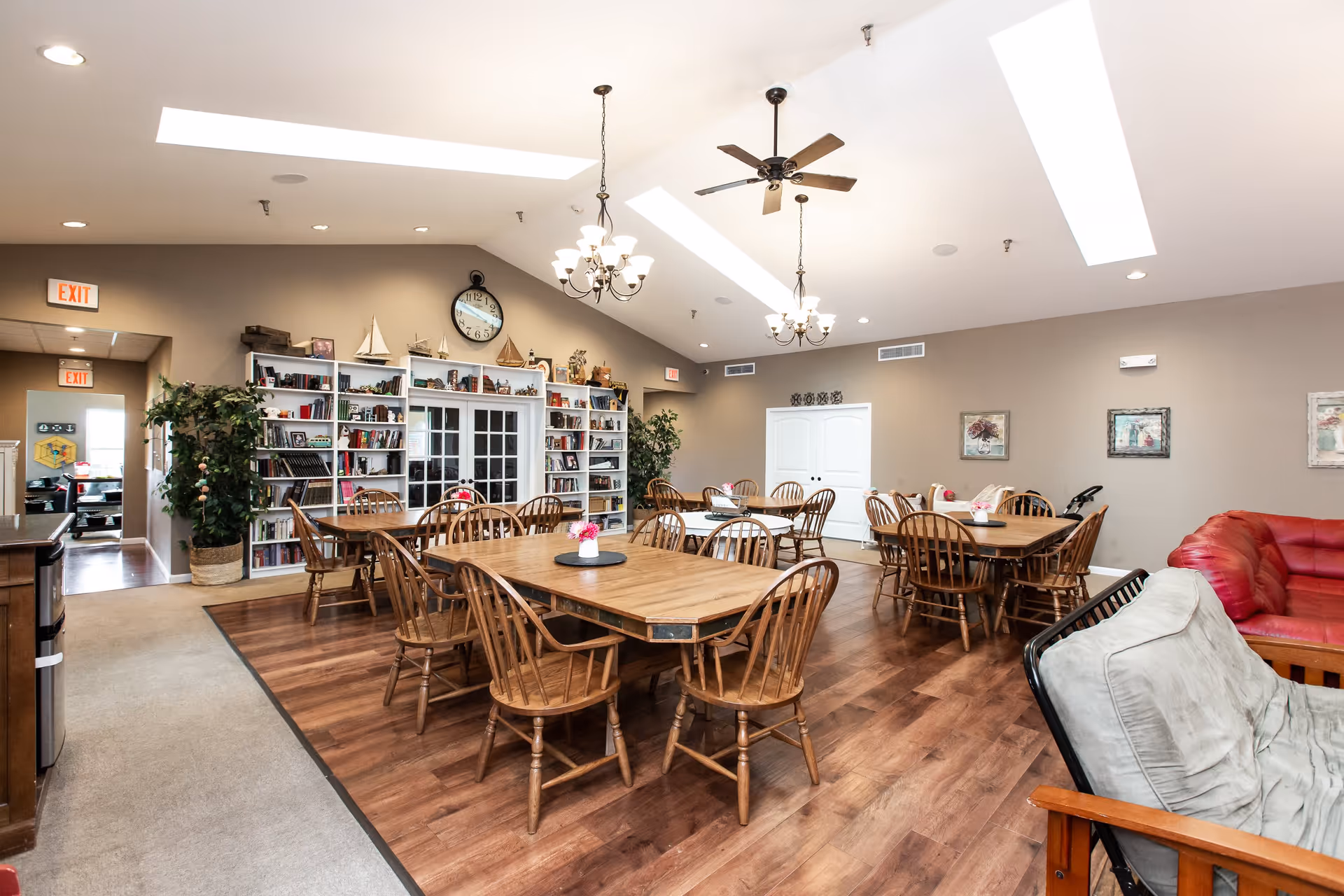 Bright communal dining and lounge area with multiple wooden tables and chairs, bookshelves, chandeliers, and seating.