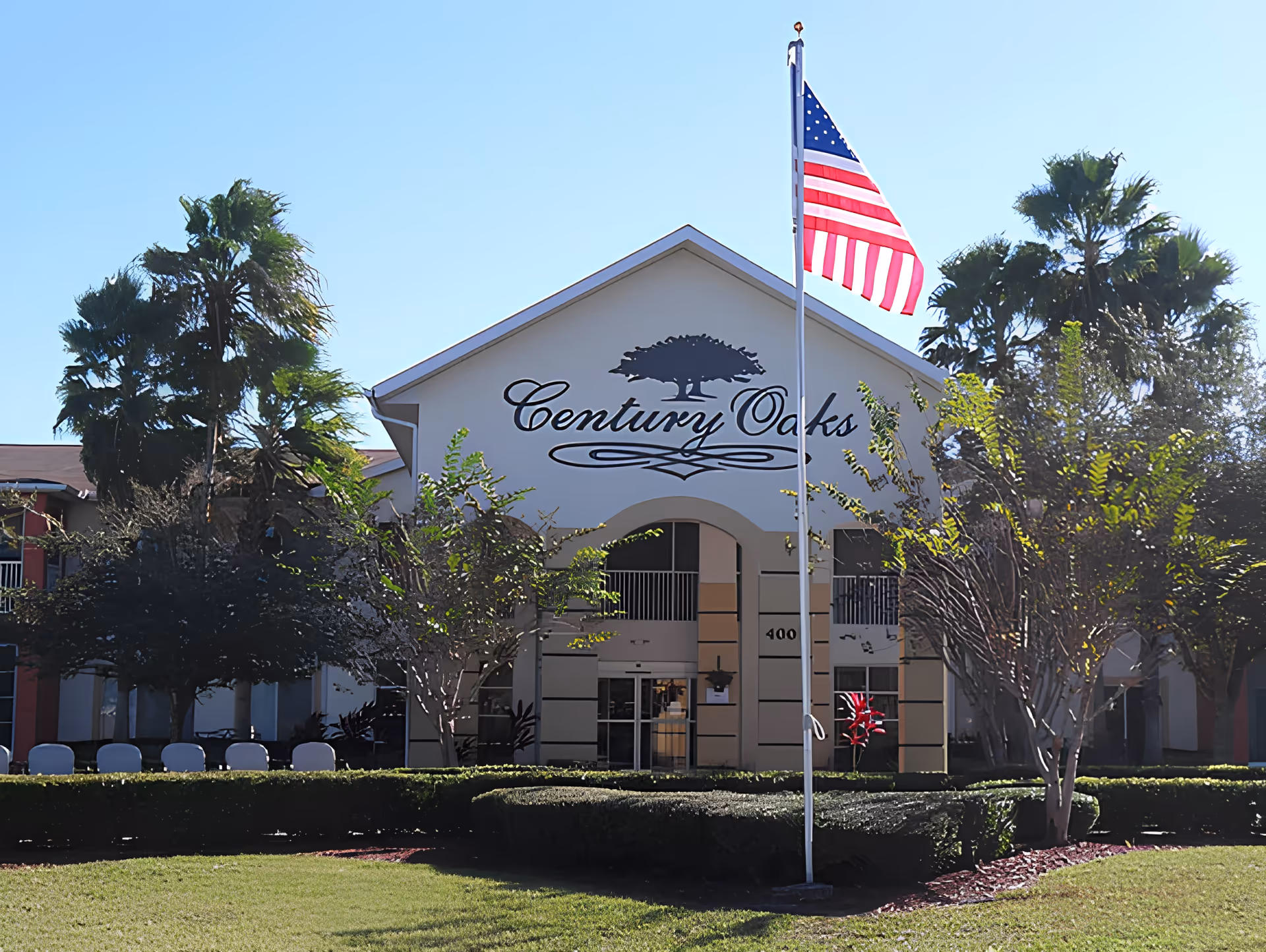 Front exterior view of the Century Oaks building with an American flag on a flagpole in front, surrounded by trees and well-maintained landscaping under a clear blue sky.