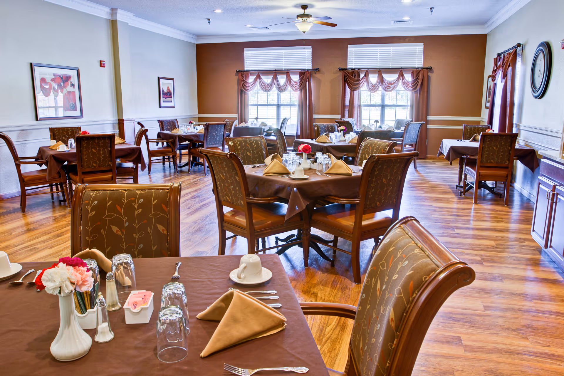 A dining room with multiple tables covered in brown tablecloths, each set with napkins, cups, glasses, and silverware. The room has wooden floors, large windows with brown curtains, framed artwork on the walls, and a ceiling fan.
