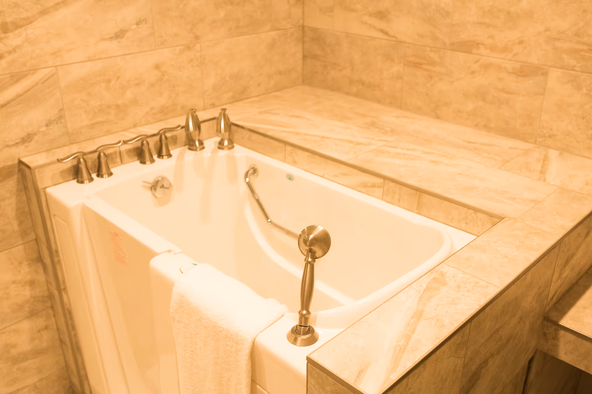 A walk-in bathtub with a white towel hanging on the side, surrounded by beige marble tiles. The tub has multiple silver faucets and a safety grab bar inside.