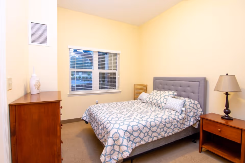 A bedroom with a bed covered in a patterned blue and white bedspread, two pillows, a wooden nightstand with a lamp, a wooden dresser with a decorative vase, a wooden chair, and a window with white blinds.