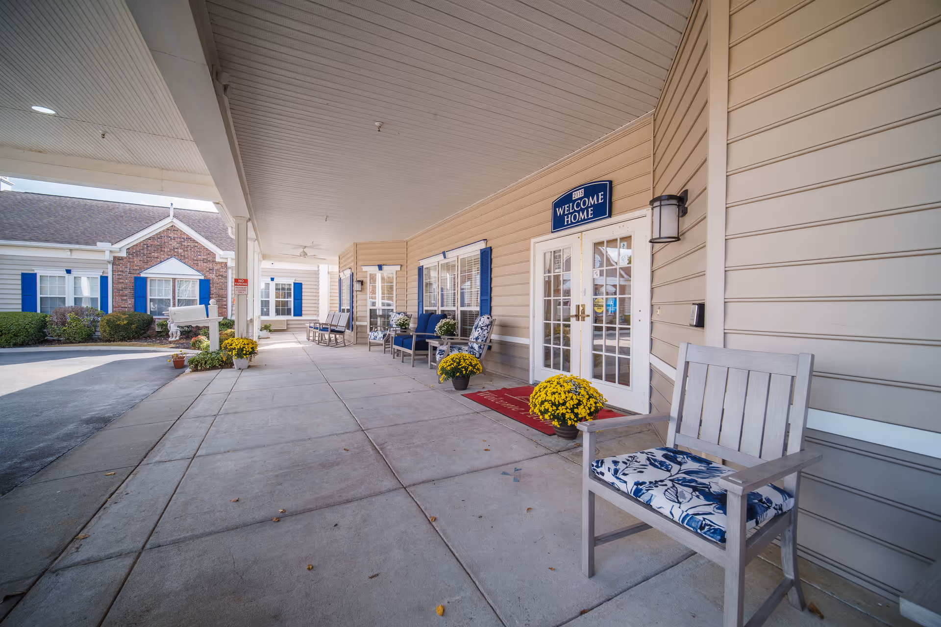Covered entrance area of a senior living facility with beige siding and blue shutters. Several chairs with floral cushions are arranged along the wall, and yellow potted flowers are placed near the entrance. A sign above the double glass doors reads 'WELCOME HOME'.