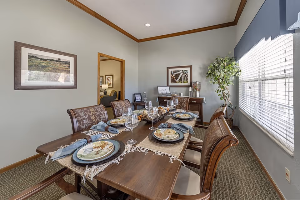 A dining room with a wooden table set for six people, featuring plates, napkins, and wine glasses. The room has light gray walls with wooden trim, a large window with blinds on the right, a framed picture on the left wall, and a potted plant in the corner near a small sideboard with decorative items.