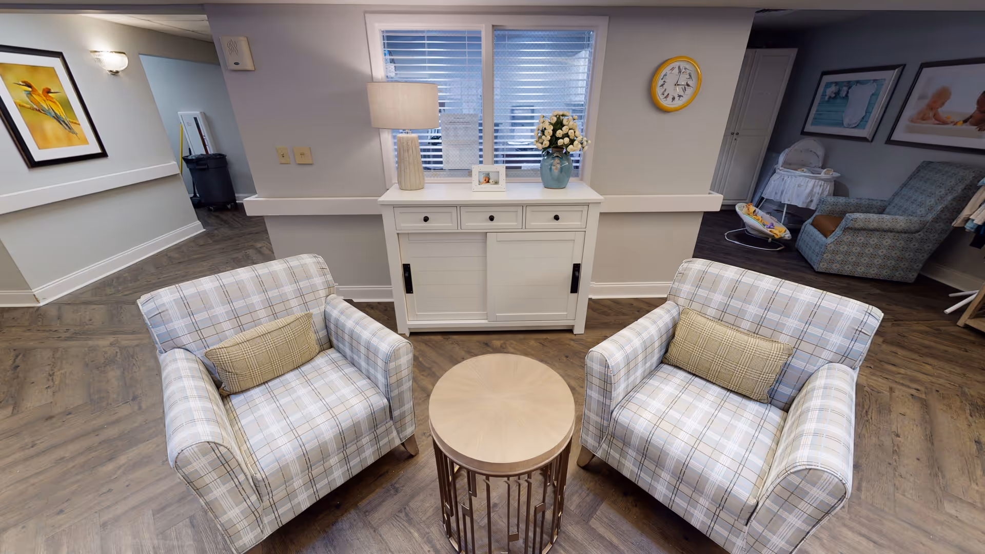 Two plaid armchairs with a round wooden side table face a white console table beneath a window in a cozy seating area.