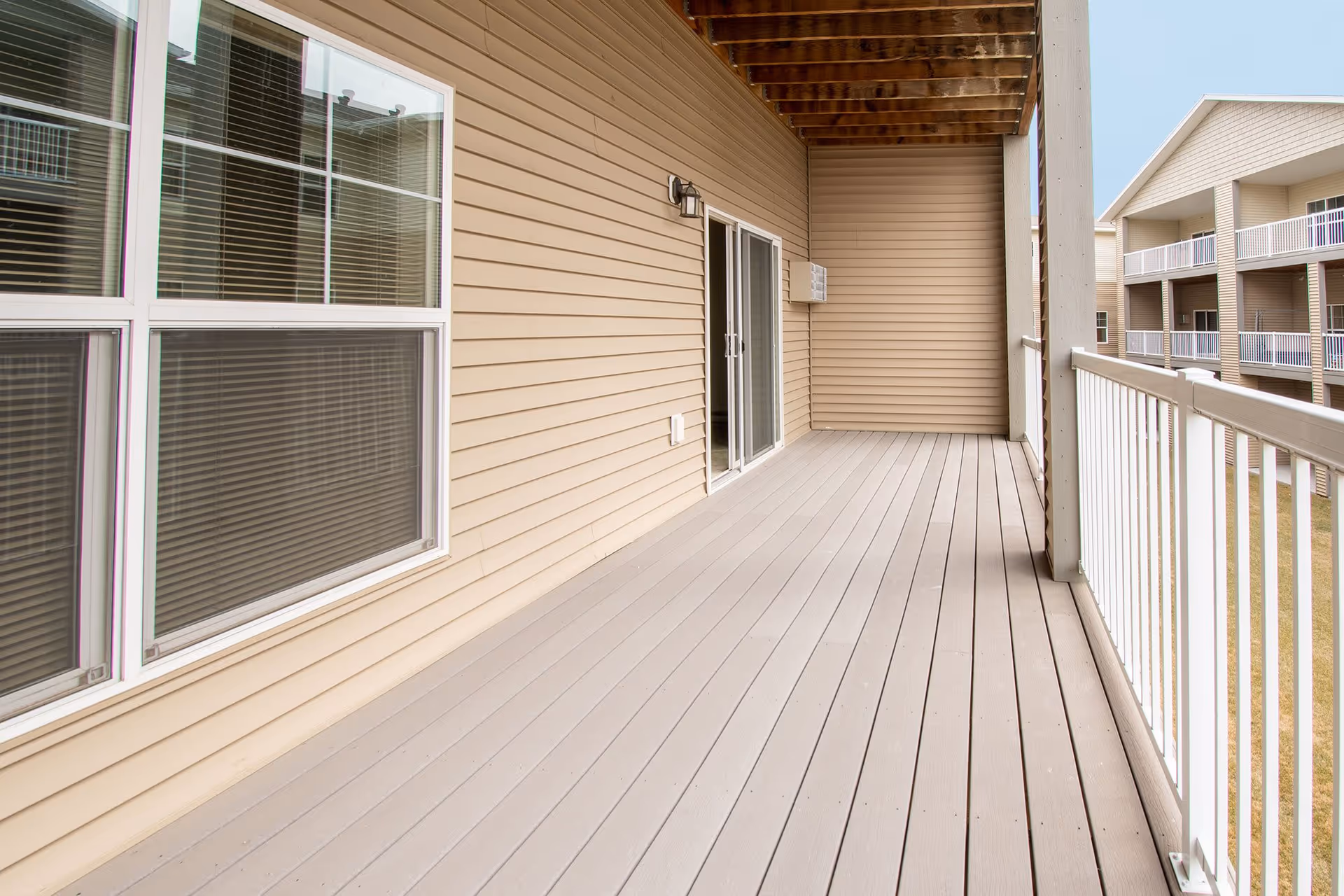 Covered second-floor balcony with beige siding, sliding glass door, windows, and white railing overlooking neighboring units.