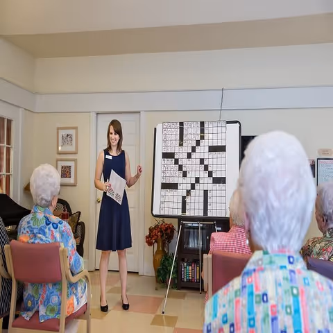 A woman in a navy blue dress stands in front of a large crossword puzzle on an easel, engaging a group of elderly people seated in chairs in a room with light-colored walls and tiled floor.