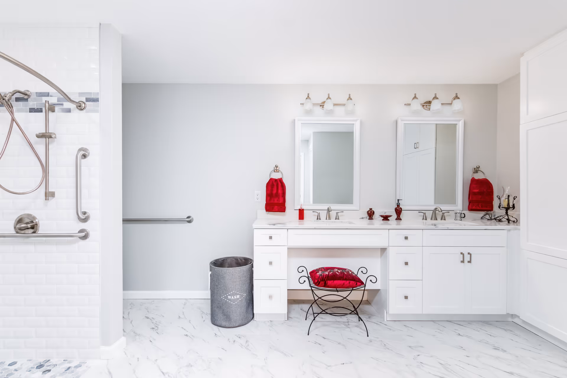 Bright and modern bathroom with white cabinetry and marble floor tiles. There are two sinks with mirrors above them, each with a set of three light fixtures. Red towels and decorative items add a pop of color. A metal chair with a red cushion is placed in front of the vanity. On the left side, there is a walk-in shower with grab bars and a handheld showerhead. A gray laundry basket labeled 'WASH' is next to the vanity.