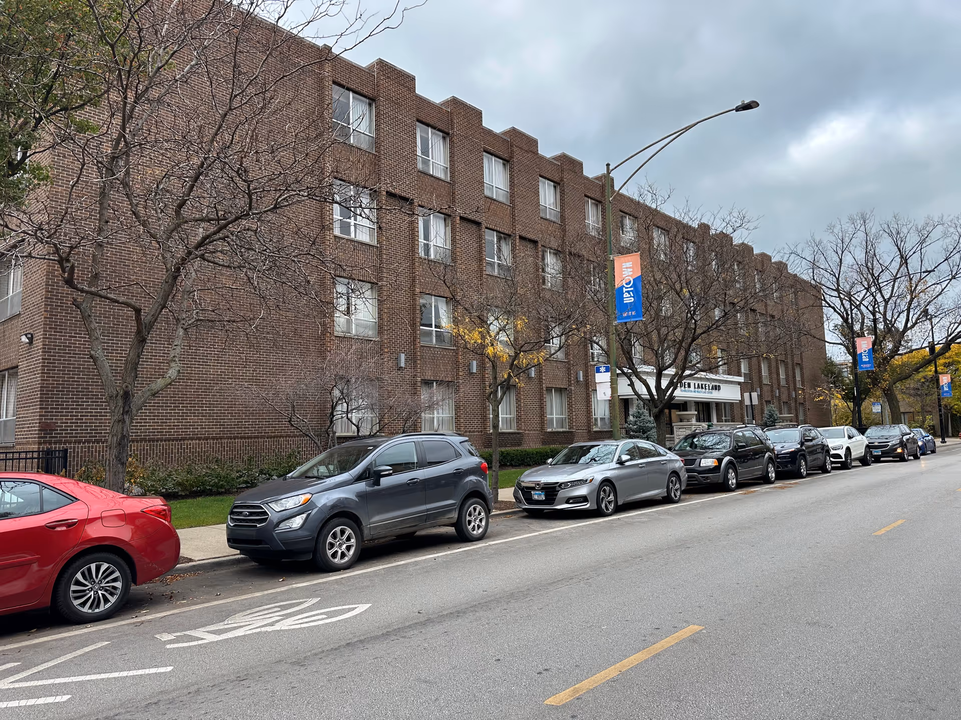 Street view of a multi-story brick building with several windows and a row of parked cars along the curb. Leafless trees line the sidewalk in front of the building under a cloudy sky. A sign on the building reads Alden Lakeland.