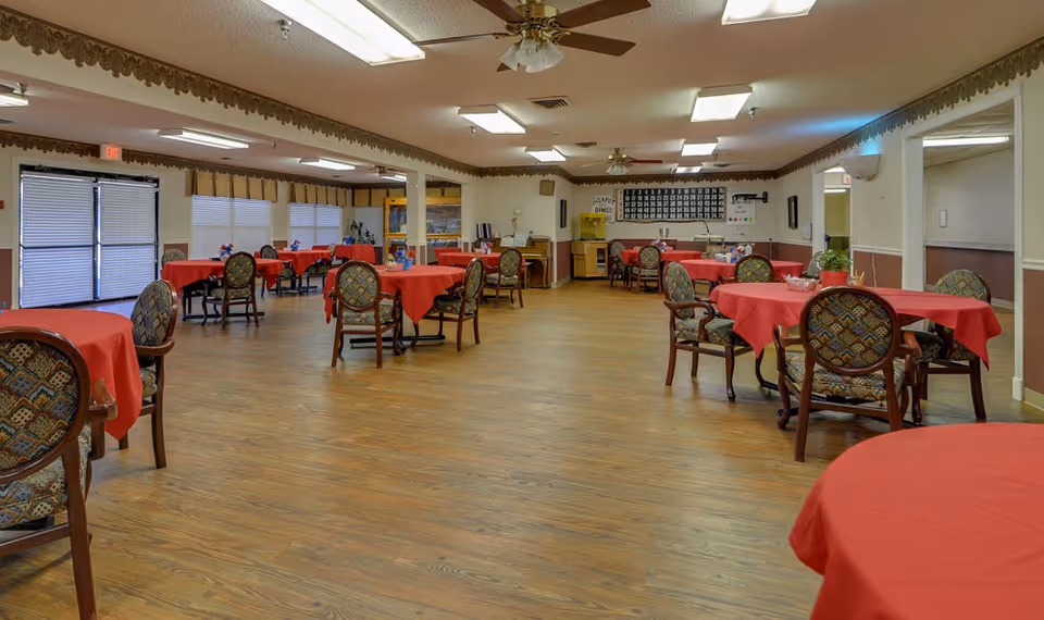 Large dining/activity room with round tables covered in red tablecloths and patterned chairs on a wood floor.