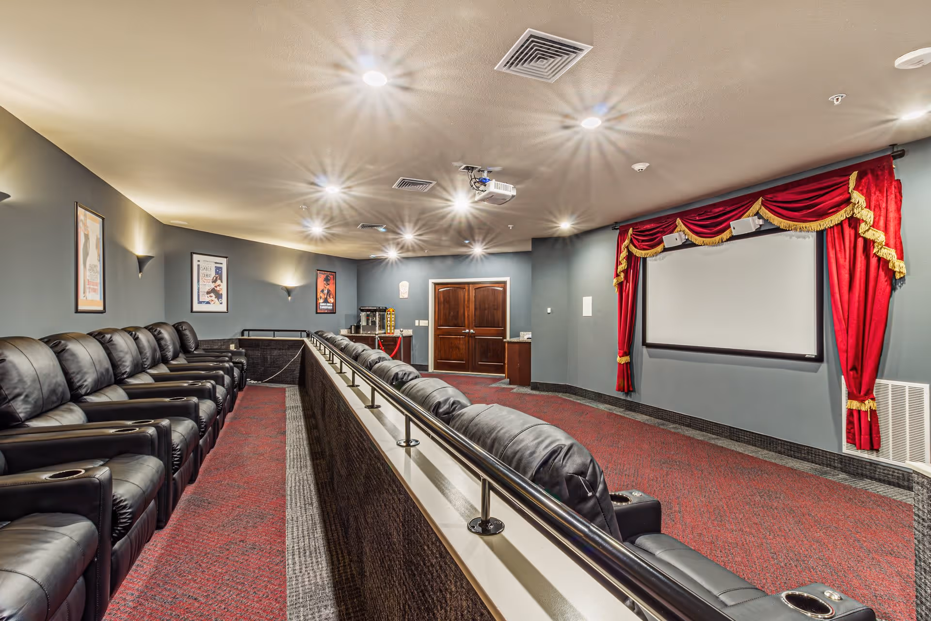 Interior view of a small theater room with two rows of black leather recliner chairs facing a large white projection screen framed by red curtains with gold trim. The room has gray walls with movie posters, recessed ceiling lights, and a popcorn machine near double wooden doors at the back.