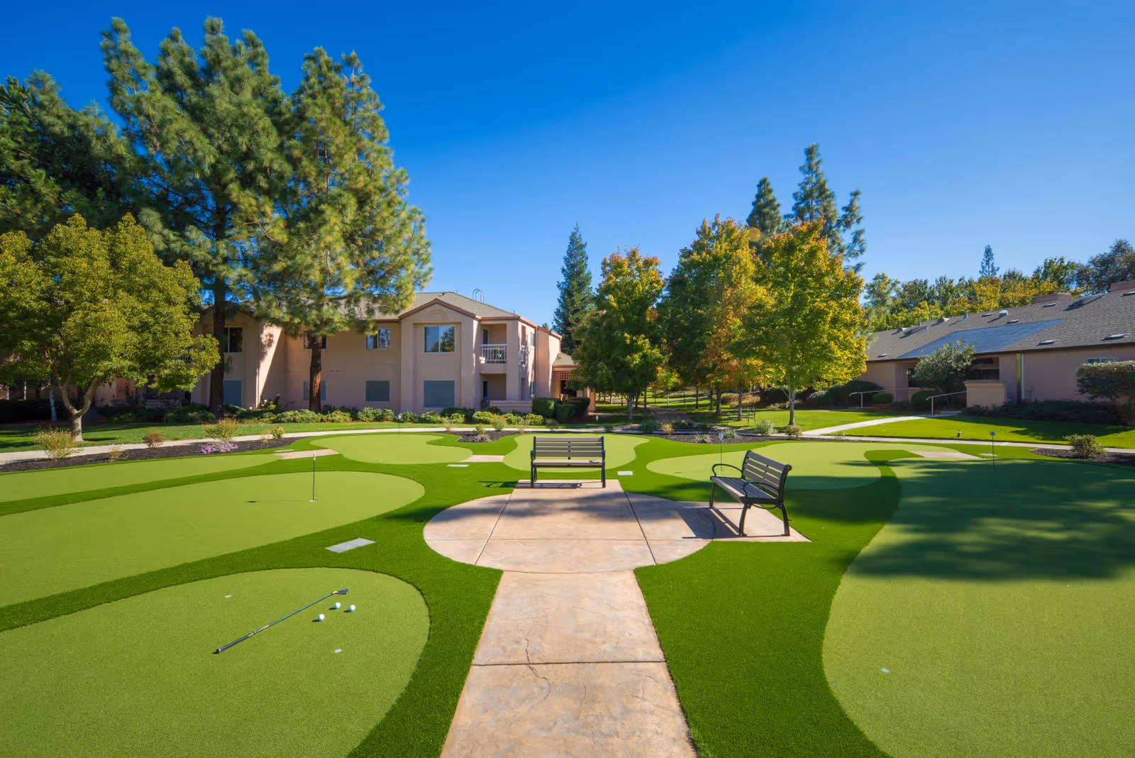 Outdoor putting green courtyard with benches, trees, and apartment-style senior living buildings under a clear blue sky.