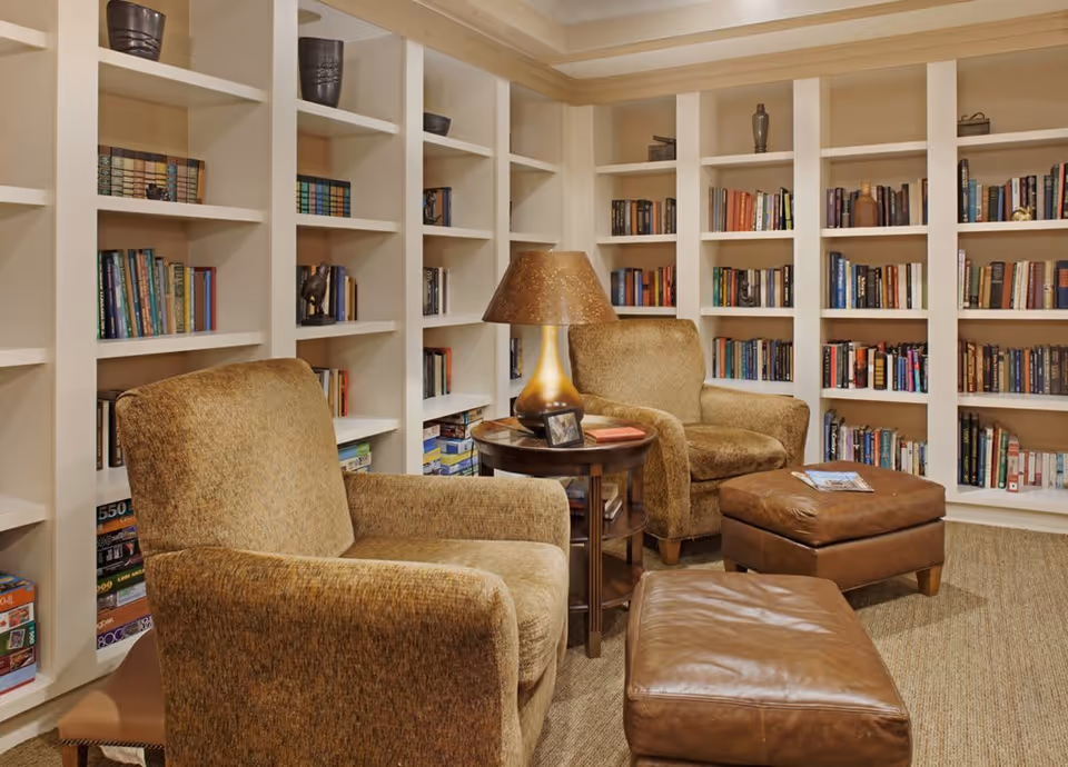 Cozy reading nook in a senior living facility with two upholstered armchairs, two leather ottomans, a round wooden side table with a lamp, and built-in bookshelves filled with books and board games.