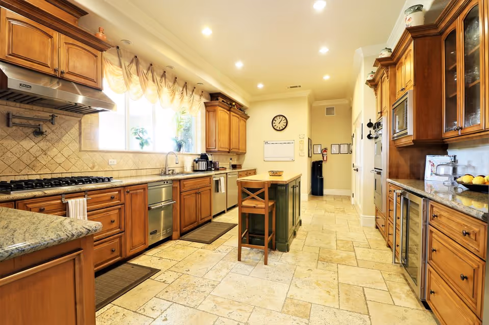 A spacious, well-lit kitchen with wooden cabinets, granite countertops, and tiled flooring. The kitchen features a large window with sheer curtains, a central island with a wooden chair, stainless steel appliances including a stove, dishwasher, and microwave, and a wall clock above a whiteboard calendar.