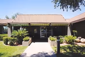Exterior view of a single-story building with a brown tiled roof and a covered entrance. The entrance is flanked by small landscaped areas with green shrubs and palm plants. A concrete pathway leads to the glass door entrance under a yellow and brown striped awning. Trees and a clear sky are visible in the background.