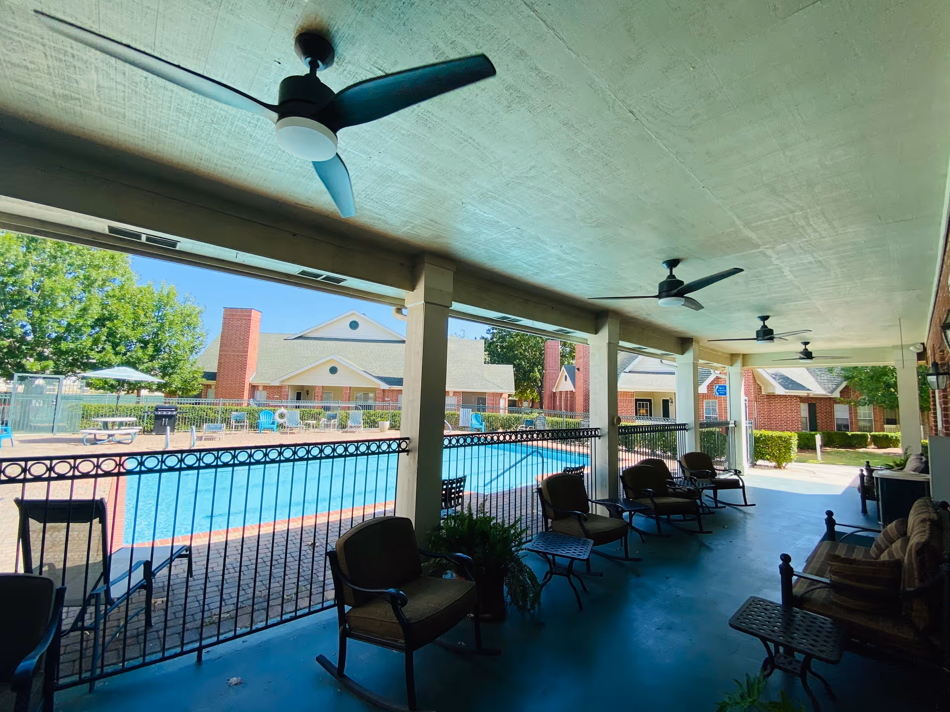 Covered patio area with ceiling fans and cushioned chairs overlooking a fenced swimming pool. In the background, there are brick buildings with white trim and green trees under a clear blue sky.