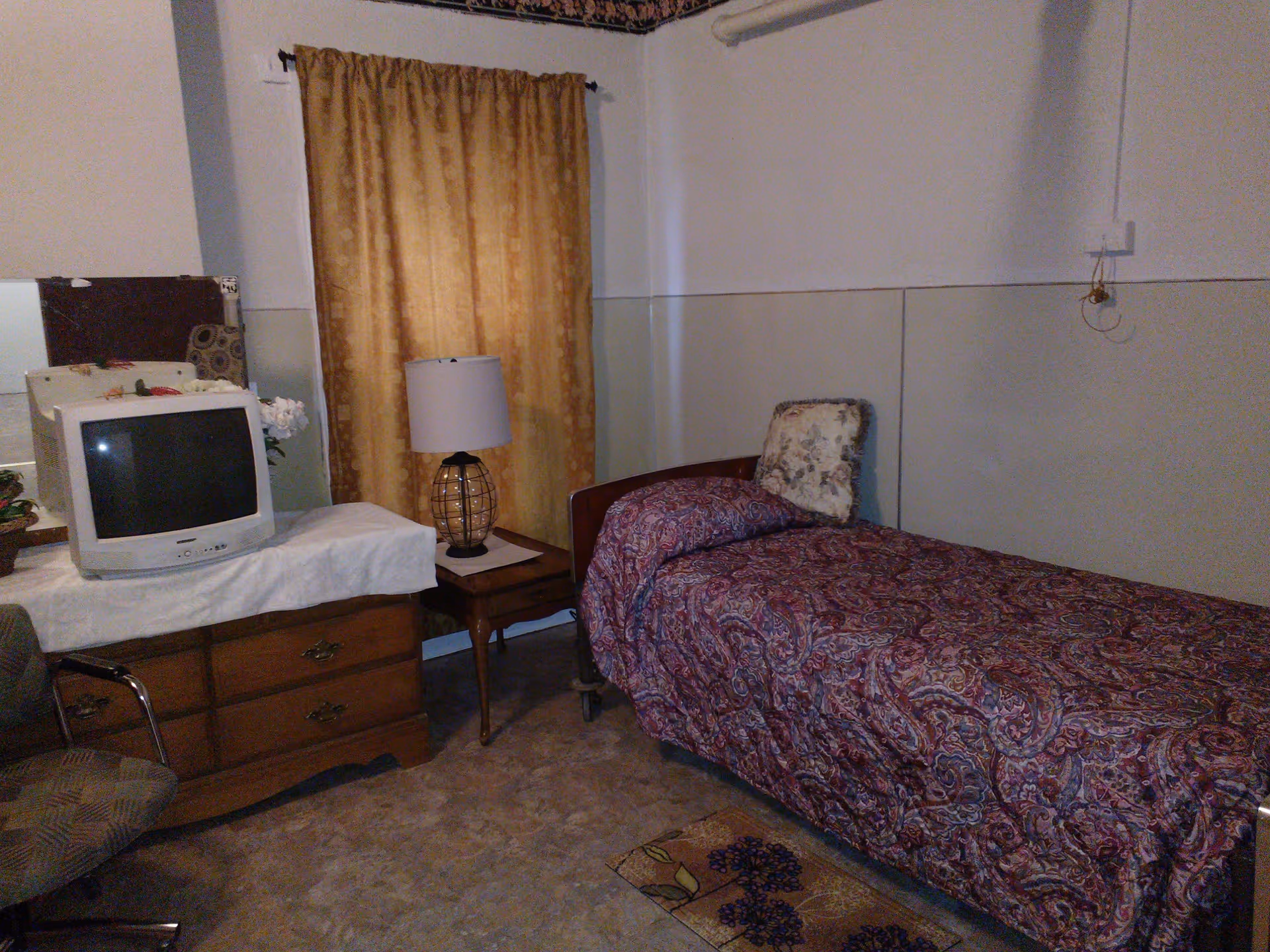 A small bedroom with a single bed covered in a patterned purple bedspread and a floral pillow. Next to the bed is a wooden nightstand with a lamp that has a white shade and a black wire base. Across from the bed is a wooden dresser with a white cloth on top, holding an old-style CRT television and a small flower arrangement. A chair with a patterned cushion is partially visible in the corner. The room has beige walls with a yellow curtain covering the window.