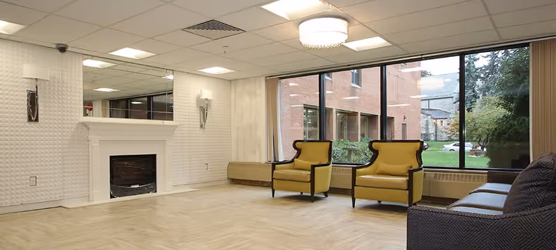 A spacious, well-lit common area with a white fireplace, two yellow armchairs, a dark gray sofa, large windows showing an outdoor view of a brick building and greenery, and a light wood floor.