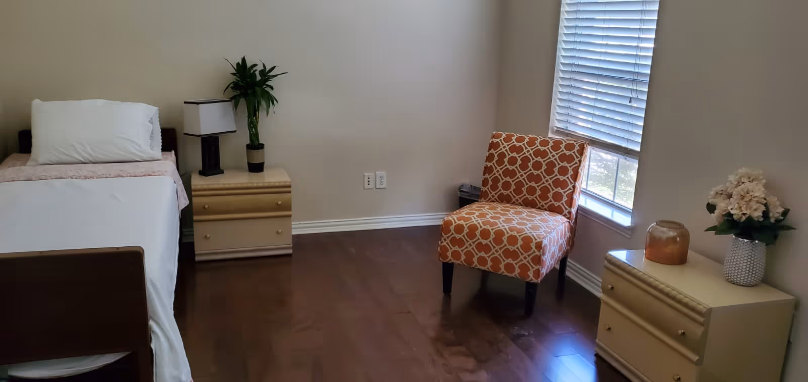 A simple bedroom with a single bed on the left side, a nightstand with a lamp and a plant next to the bed, an orange patterned chair near a window with blinds, and a dresser with a vase of flowers and a decorative item on the right side. The room has wooden flooring and light-colored walls.