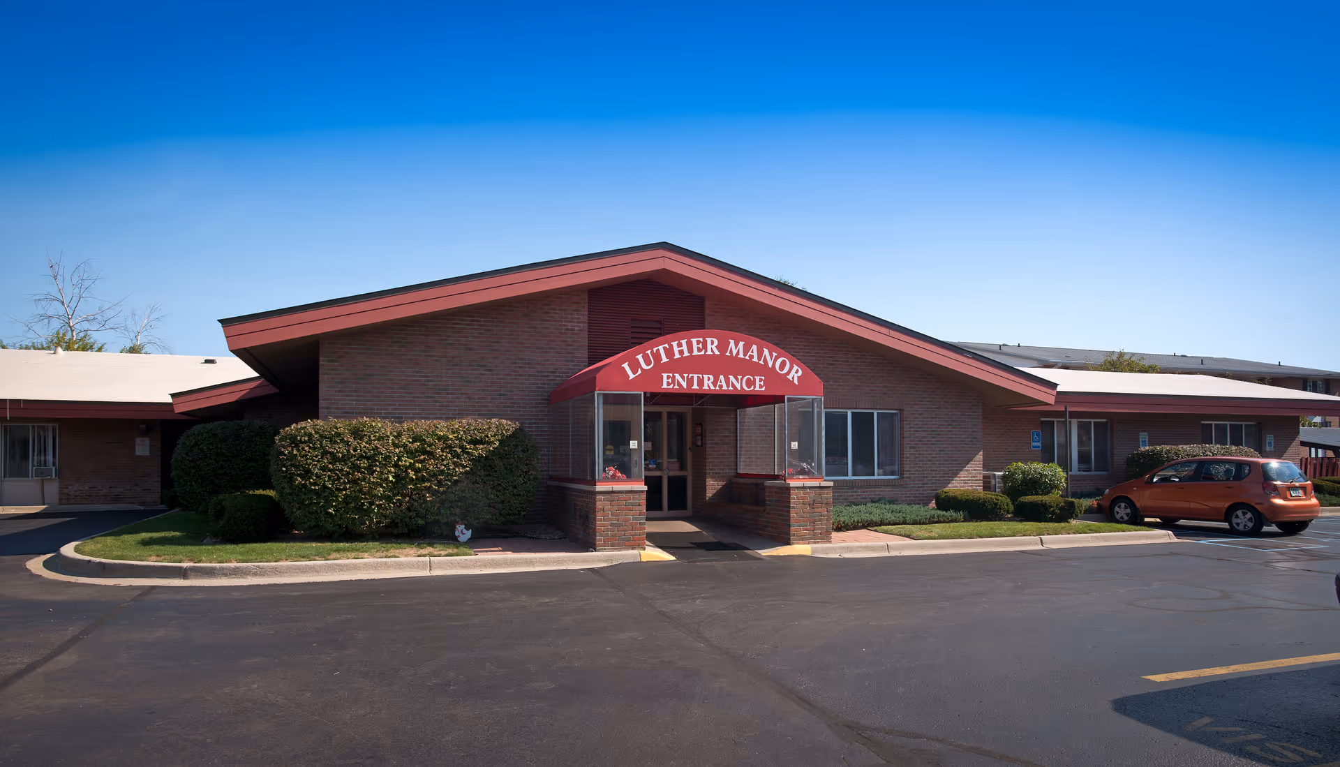Single-story brick facility front with a red entrance awning reading 'LUTHER MANOR ENTRANCE' and a small parking area.