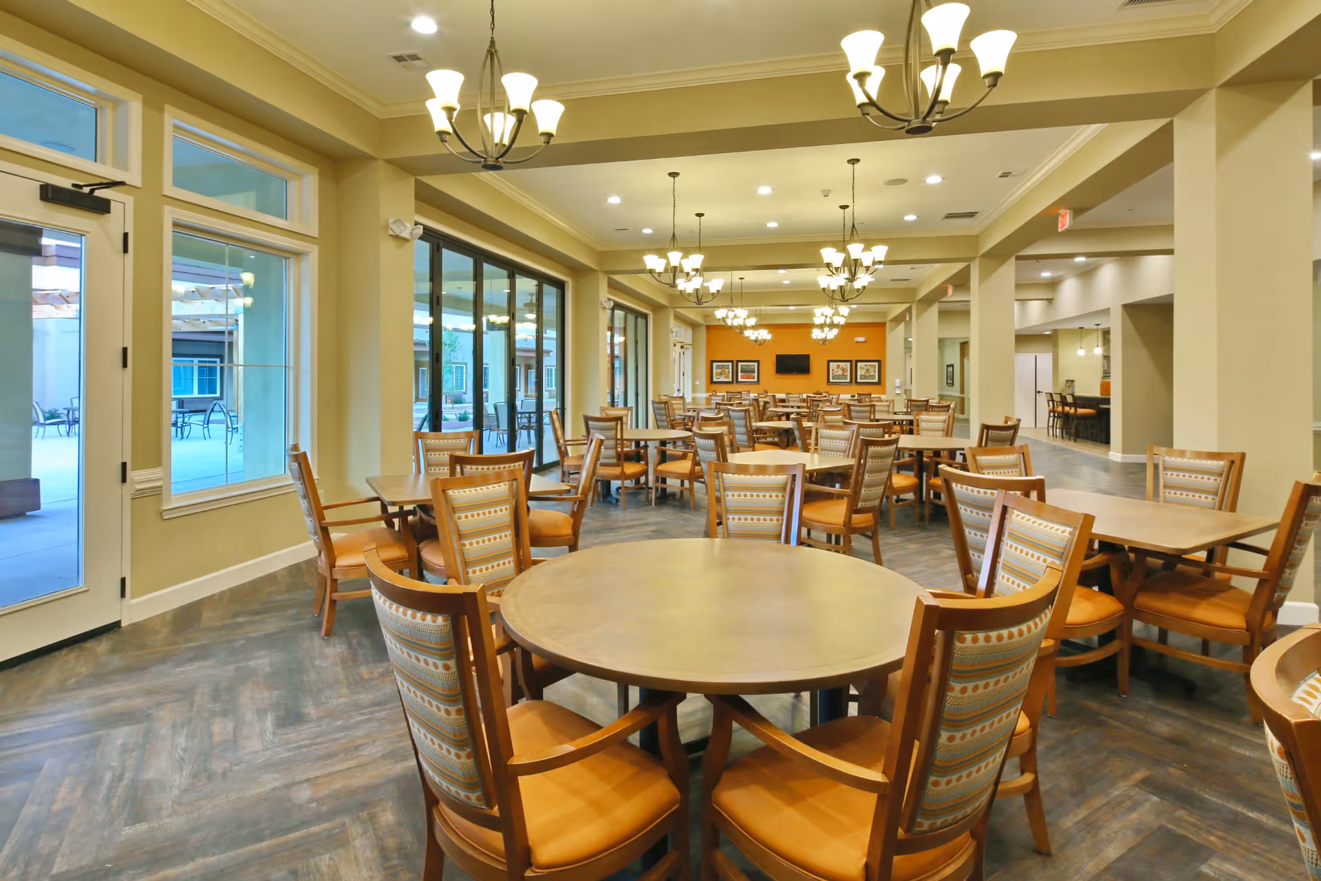 A spacious dining room with multiple round and rectangular wooden tables surrounded by cushioned chairs with patterned backs. The room features large windows and glass doors on one side, allowing natural light to enter and providing a view of an outdoor patio area. Several chandeliers hang from the ceiling, illuminating the room. The walls are painted in neutral tones with an accent orange wall at the far end, decorated with framed artwork and a mounted flat-screen TV.