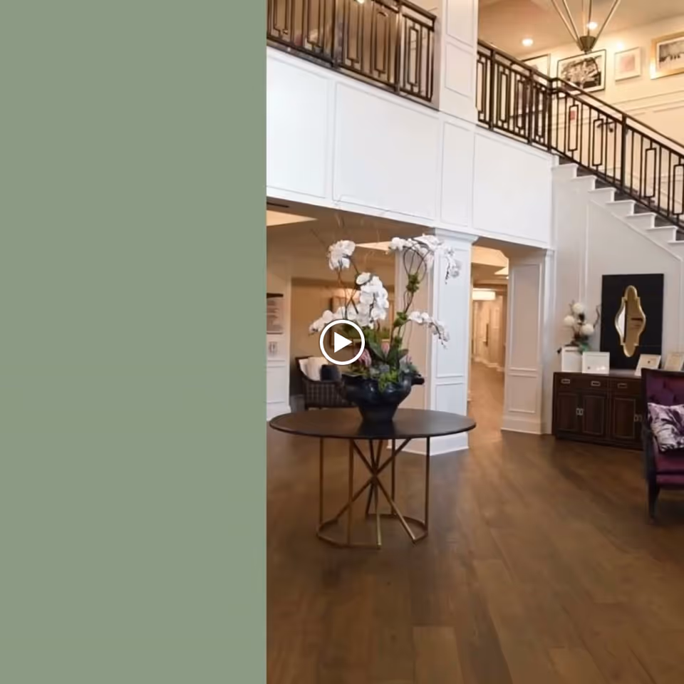 Interior view of a senior living facility lobby with a round table holding a large floral arrangement in the center. The space features wooden flooring, white paneled walls, a staircase with a black railing, and framed artwork on the walls. There is a purple armchair and a wooden cabinet with decorative items on the right side.