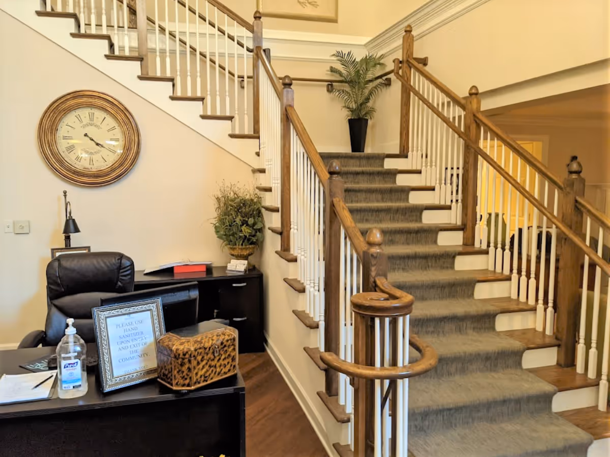 Interior view of a senior living facility reception area with a wooden staircase carpeted in gray leading to the upper floor. A black office chair and desk with hand sanitizer, a framed sign, and a decorative box are visible. A large round wall clock hangs on the wall above a small table with a plant. The walls are painted light beige.