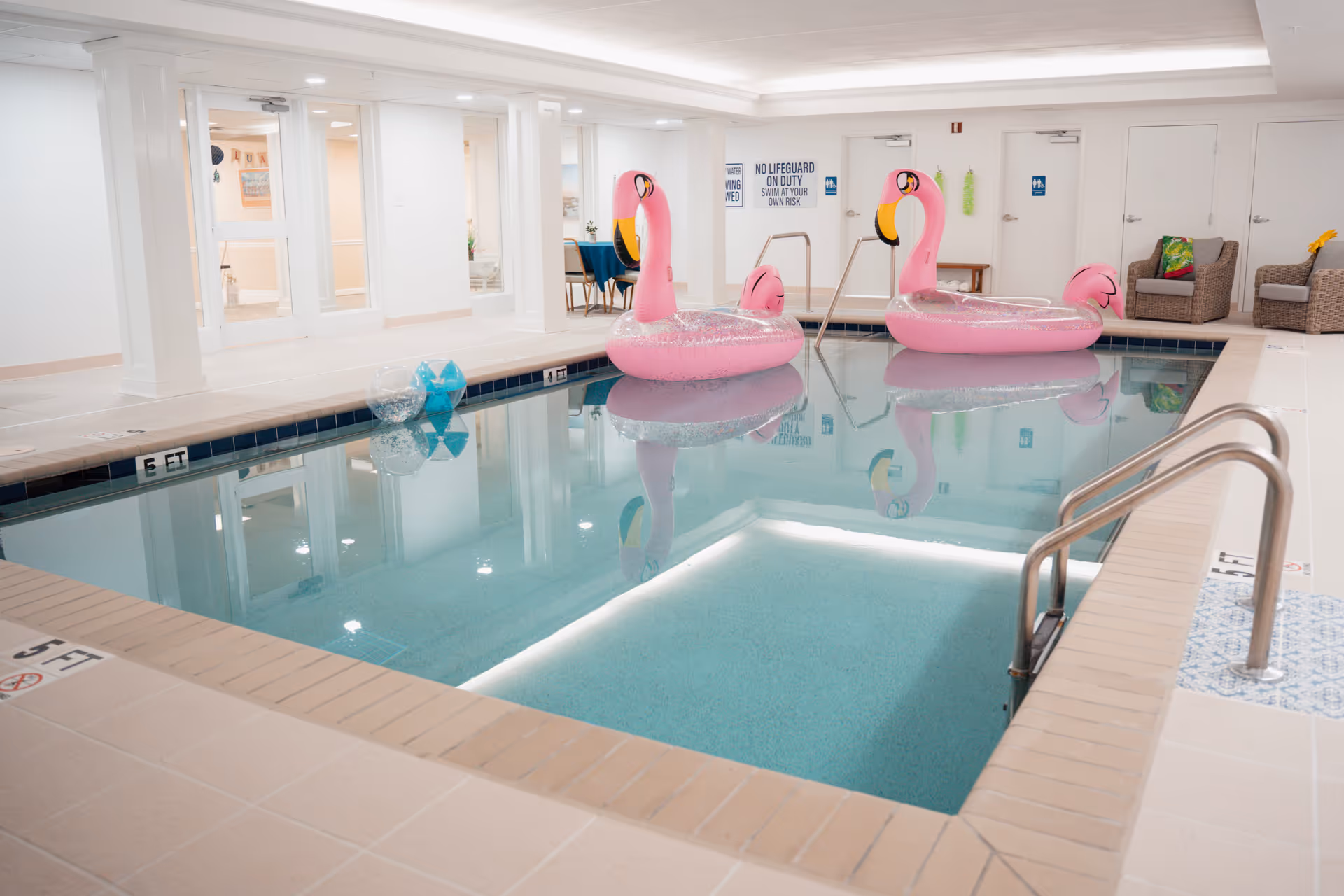 Indoor swimming pool with two large pink flamingo floaties in the water. The pool area has beige tiled flooring and white walls with several doors and chairs along the side. Signs on the wall indicate no lifeguard on duty and restroom locations.