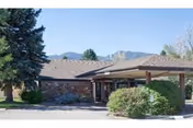 Low single-story brick building entrance with a covered porte-cochere, shrubs, trees, and mountains in the background.