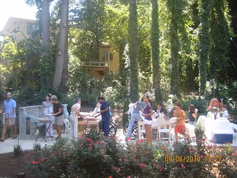 Residents and staff gathered around tables in a shaded outdoor courtyard with trees and flower beds.