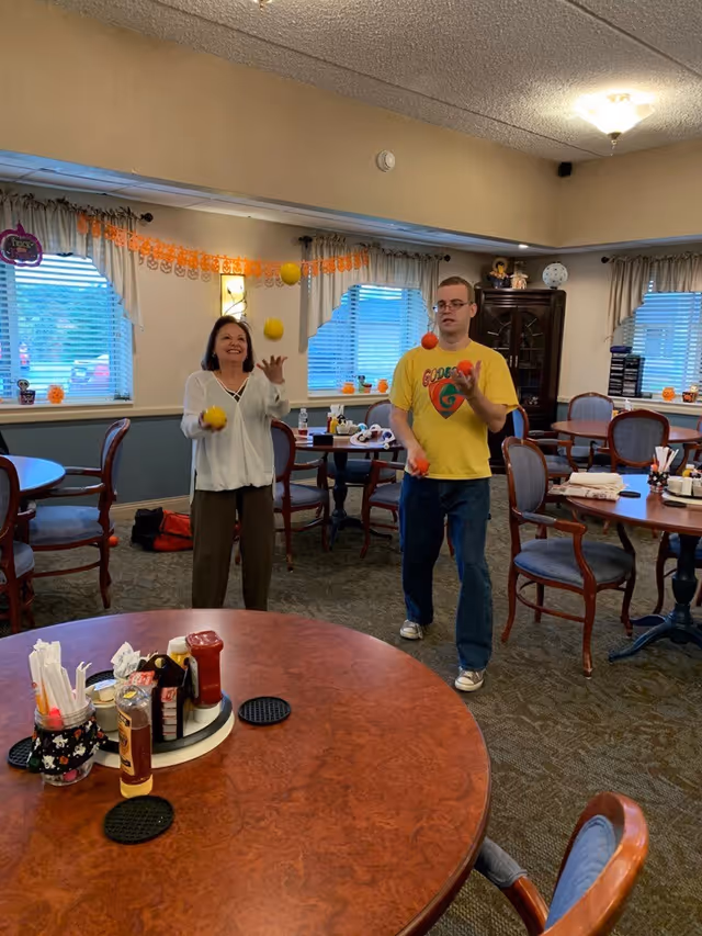 Two people juggling colorful balls in a decorated dining/activity room with tables and chairs.