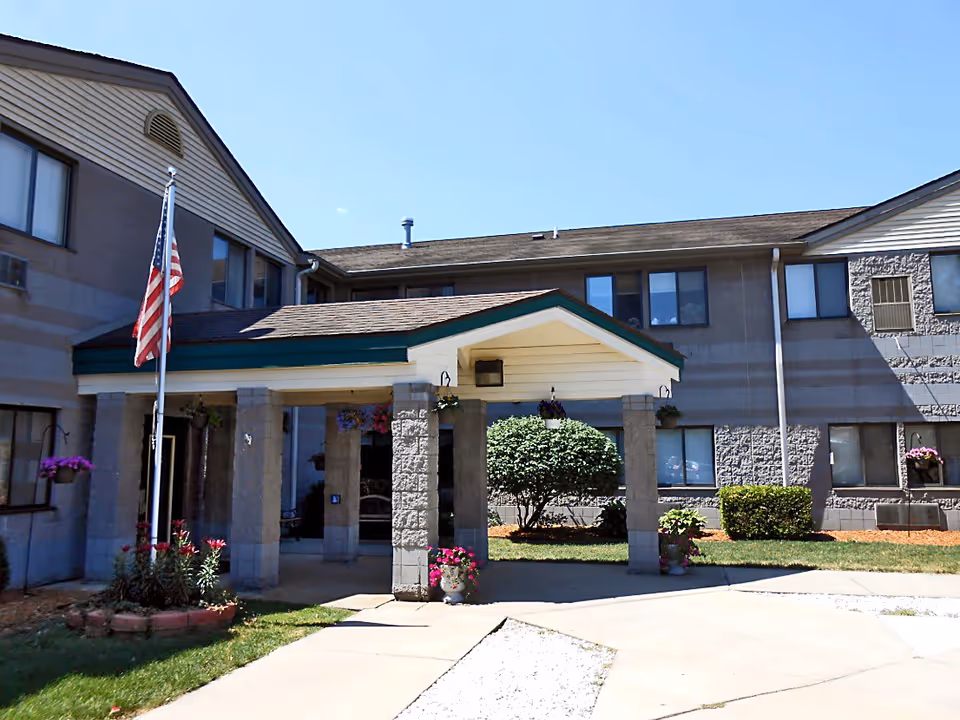 Exterior view of Wood Ridge Assisted Living facility showing the entrance with a covered porch supported by stone pillars. There is an American flag on a flagpole to the left, flower pots with blooming flowers, and neatly trimmed bushes around the building. The sky is clear and blue.