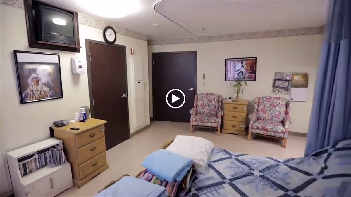 A room with a bed covered in a blue and white patterned blanket, two floral armchairs, a wooden dresser, a small bookshelf, a wall-mounted TV, and framed artwork on the walls. There are two closed doors and a blue privacy curtain partially visible on the right side.