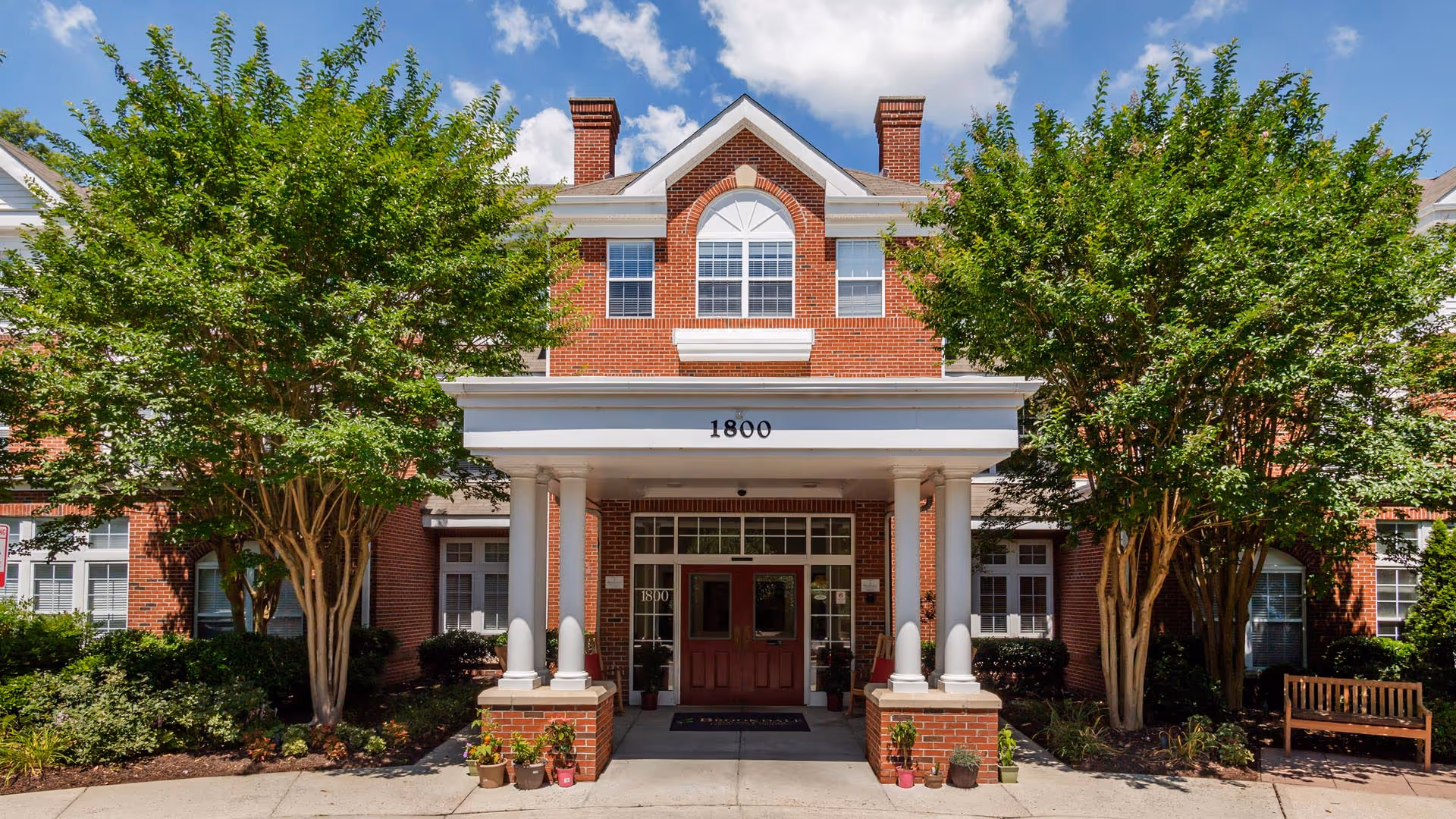Front exterior view of a brick building with white columns at the entrance, the number 1800 displayed above the doorway, surrounded by green trees and plants under a partly cloudy blue sky.