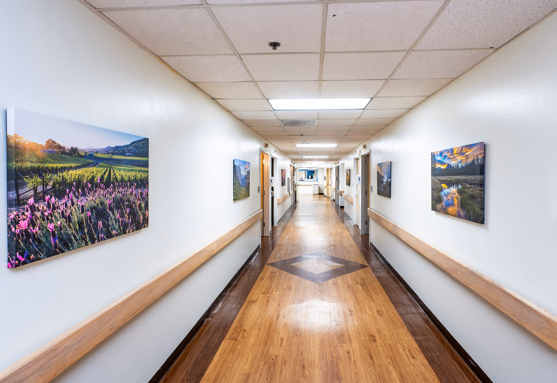 A long, well-lit hallway in a care center with wooden flooring and white walls. The walls are decorated with colorful landscape photographs. Handrails run along both sides of the hallway, and several doors are visible along the corridor.