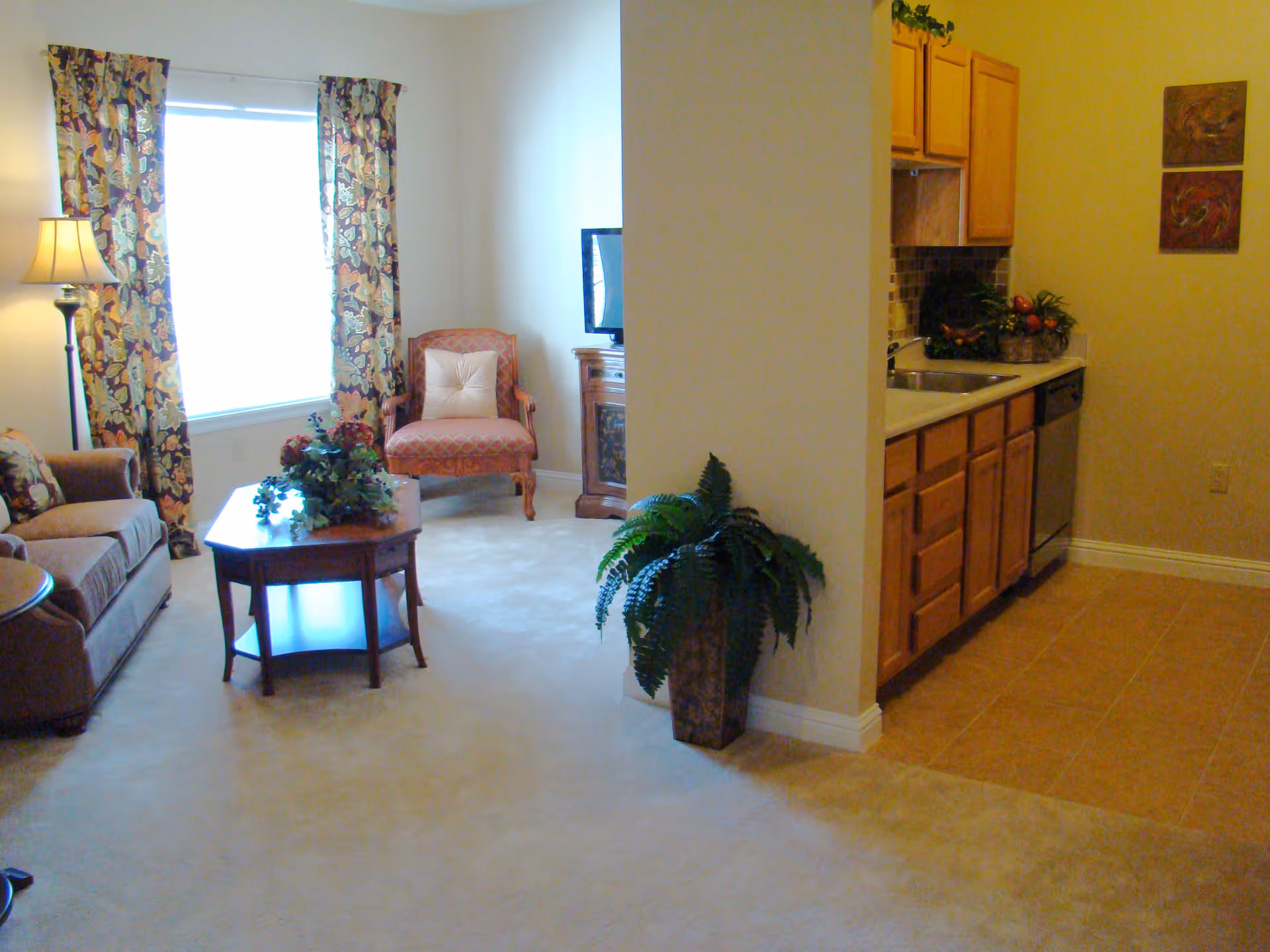 A cozy living area with a brown sofa, a wooden coffee table with a floral arrangement, a patterned armchair with a cushion, and a floor lamp next to a window with floral curtains. Adjacent to the living area is a small kitchen with wooden cabinets, a sink, a dishwasher, and a tiled floor. A potted plant is placed near the corner dividing the living room and kitchen.