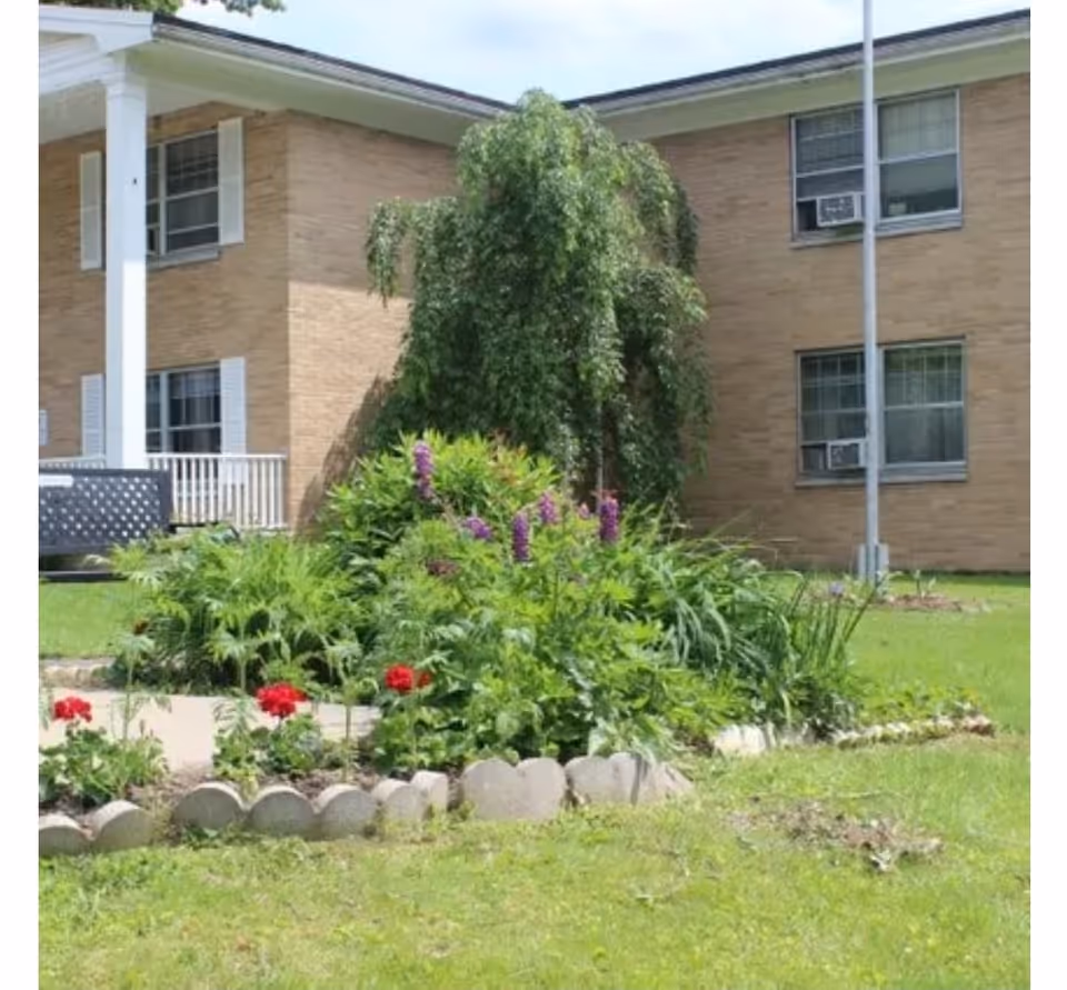 A landscaped flowerbed with purple and red blooms in front of a two-story brick building with windows and a flagpole.
