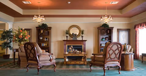 A cozy living room area with four upholstered armchairs arranged around a wooden coffee table in front of a fireplace. The fireplace has a decorative round mirror above it and two matching wooden shelves on either side with plants and decorative items. The room has green carpet, warm lighting from ceiling fixtures, and large windows with curtains letting in natural light.