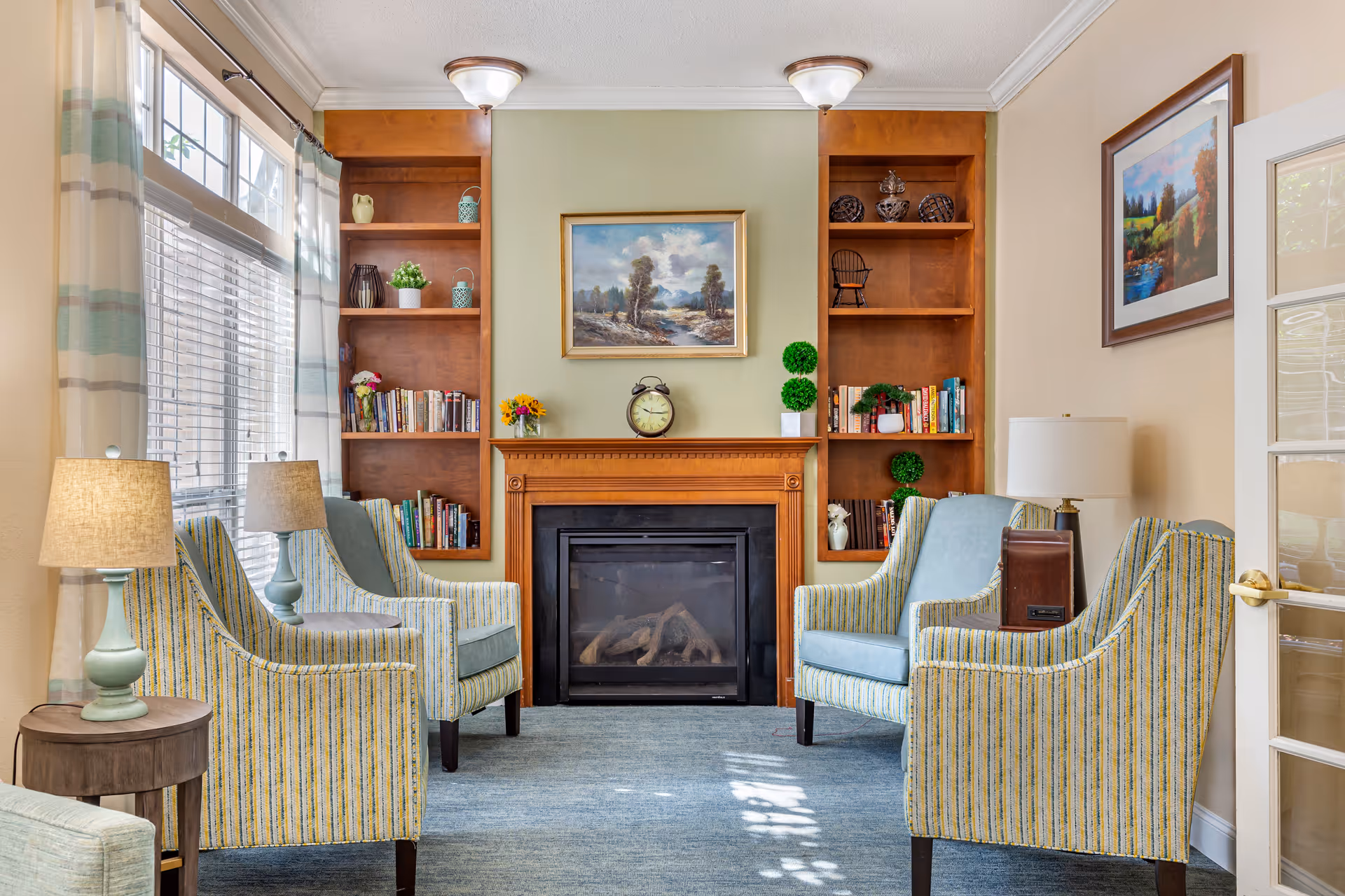 A cozy living room with four striped armchairs arranged around a fireplace. The fireplace has a wooden mantel with a clock and small plants on it. Built-in wooden shelves flank the fireplace, holding books, decorative items, and plants. Large windows with sheer curtains allow natural light to fill the room. Two table lamps with beige shades are placed on small wooden side tables next to the chairs. A framed landscape painting hangs above the fireplace, and another framed artwork is on the adjacent wall.
