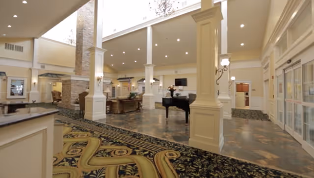 Spacious and elegant senior living facility common area with high ceilings, large white columns, a grand piano, comfortable seating, and decorative carpeting. The area is well-lit with natural light from a skylight and wall sconces, creating a welcoming atmosphere.