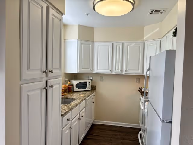 A small kitchen with white cabinets, granite countertops, a white refrigerator, a microwave, and a sink. The floor is dark wood, and there is a round ceiling light fixture.
