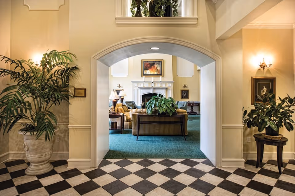 View through an arched doorway into a cozy living room with a green carpet, a fireplace, framed artwork above the mantel, and comfortable seating including sofas and armchairs. The foreground features a black and white checkered floor with large potted plants on either side of the archway, wall sconces, and soft yellow walls.