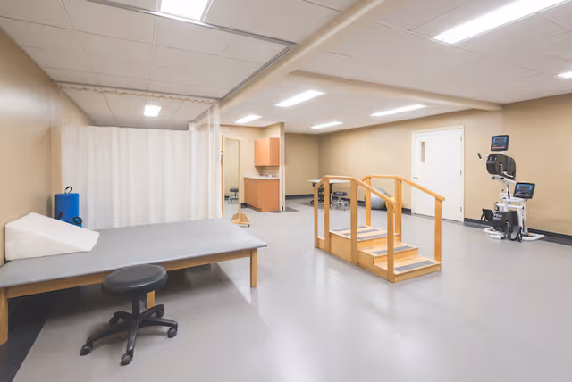 Rehabilitation therapy room with an exam table, stool, wooden practice stairs with handrails, exercise equipment, and a privacy curtain.