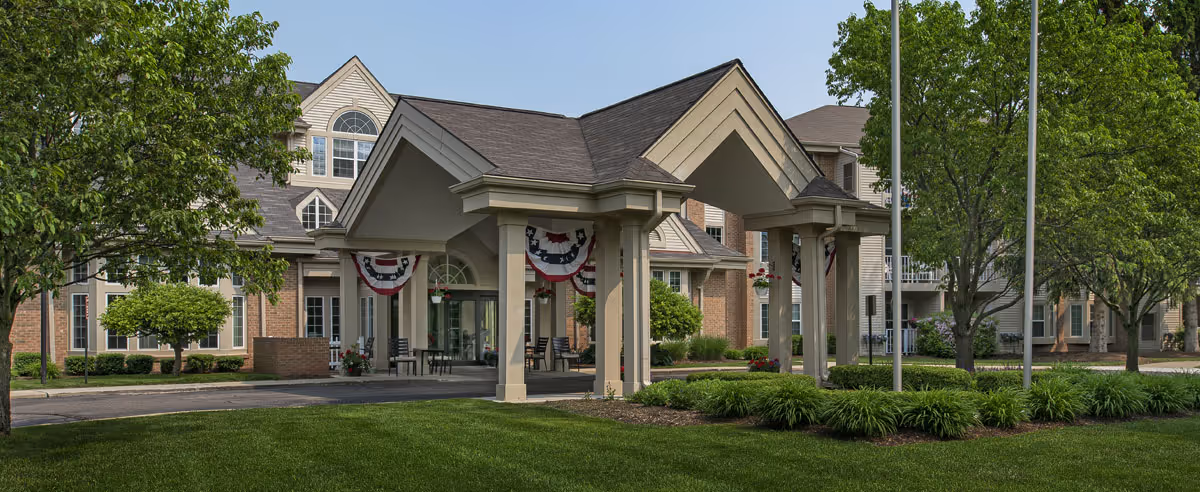 Front entrance of a senior living building with a covered porte-cochere, decorative bunting, and a manicured lawn.