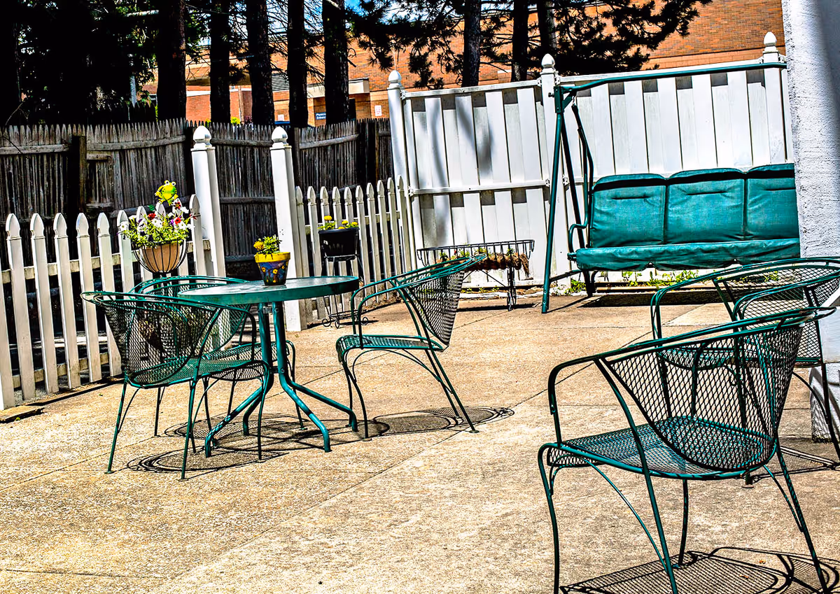 Outdoor patio area with green metal chairs and tables, a green cushioned swing, and white picket fences surrounding the space. There are potted plants on the tables and along the fence.