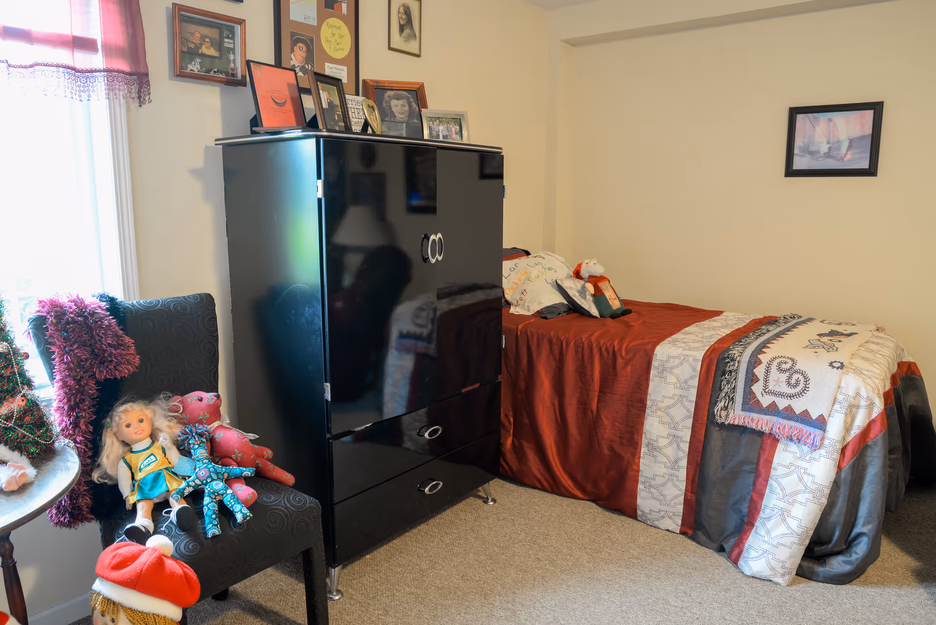 A cozy bedroom with a single bed covered in a red, white, and gray patterned quilt. Next to the bed is a black cabinet with framed photos and decorations on top. A chair near the window holds several dolls and a small Christmas tree decoration. The room has beige walls with a framed picture hanging above the bed.