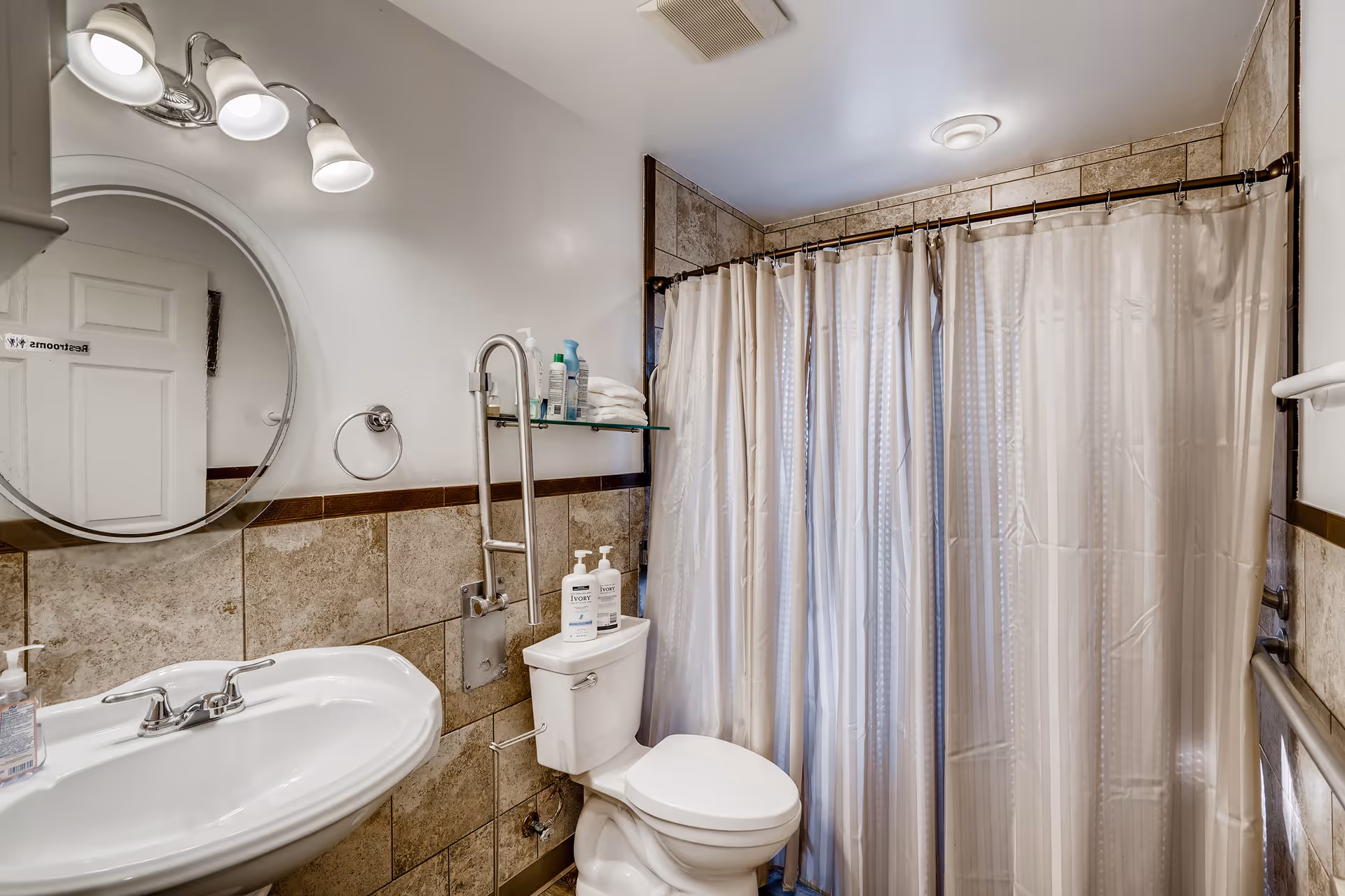 A bathroom with a white sink and faucet, a round mirror above the sink, a toilet with two bottles of Ivory soap on the tank, a glass shelf holding toiletries and towels, a shower with a beige curtain, and tiled walls with a brown trim.