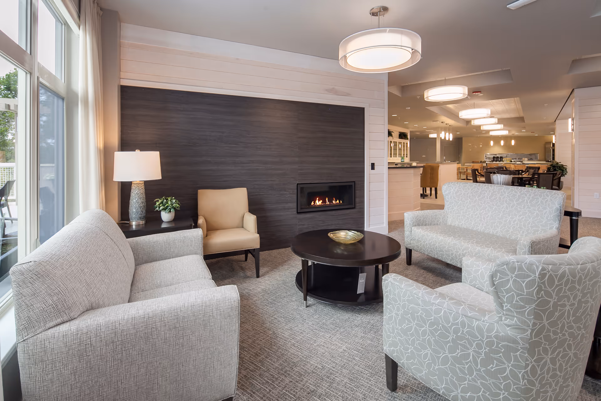A modern living room area with a light gray sofa, a beige armchair, a patterned armchair, and a matching loveseat arranged around a dark round coffee table. There is a small side table with a lamp and a plant next to the beige armchair. A sleek, built-in fireplace is set into a dark wood accent wall. Large windows with sheer curtains allow natural light to fill the room. In the background, there is a dining area with tables and chairs under pendant lighting.